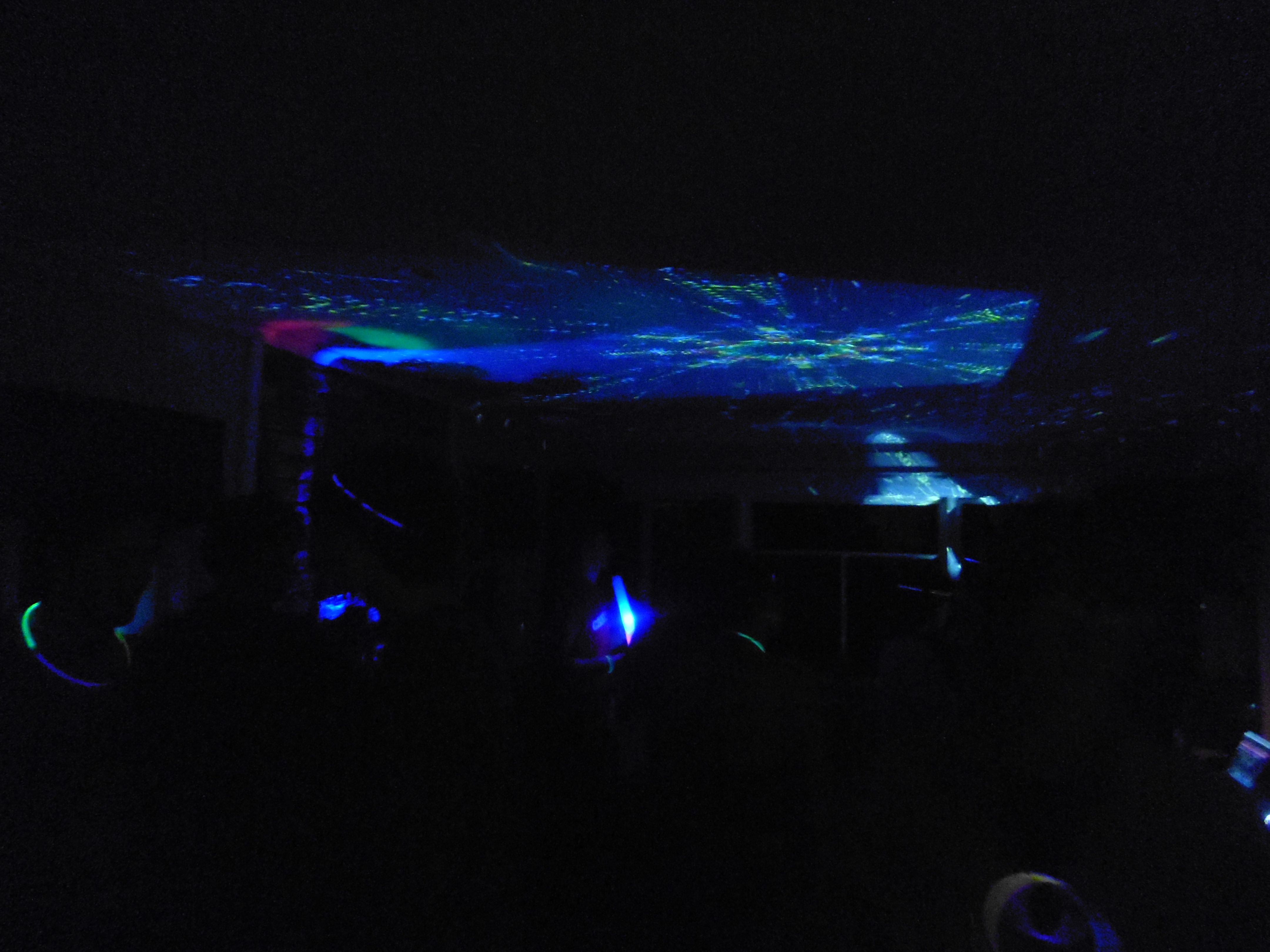 Dark room with pattern on ceiling lit by projectors. People dancing in room barely visible, with glowsticks around peoples arms and necks.